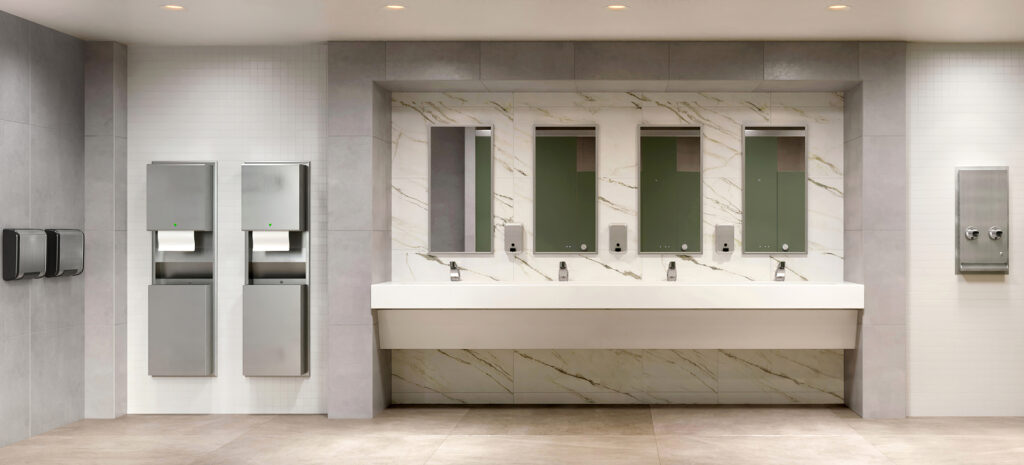 A large washroom with four individual sinks, mirrors, and soap dispensers, plus two hand dryers and roll towel dispensers and a napkin/tampon vendor.
